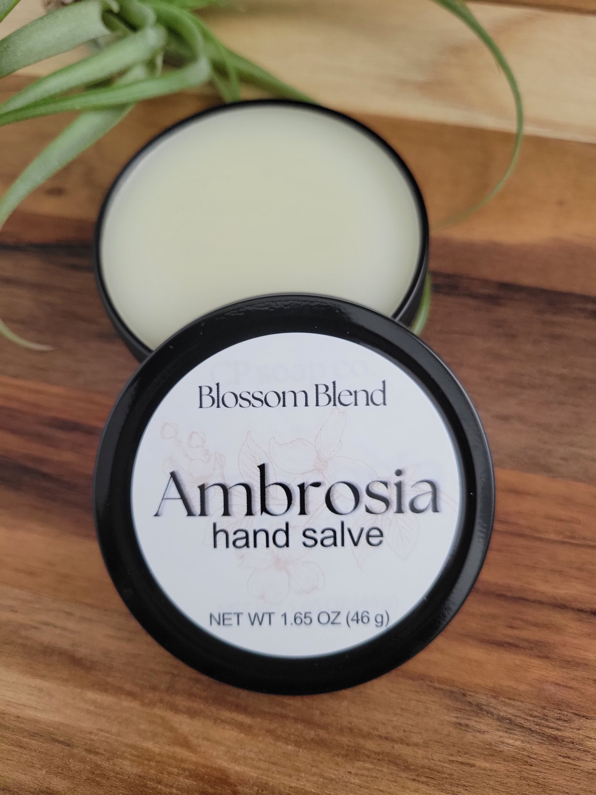 Jar of Ambrosia Blossom Blend hand salve on a wooden surface with a plant in the background.