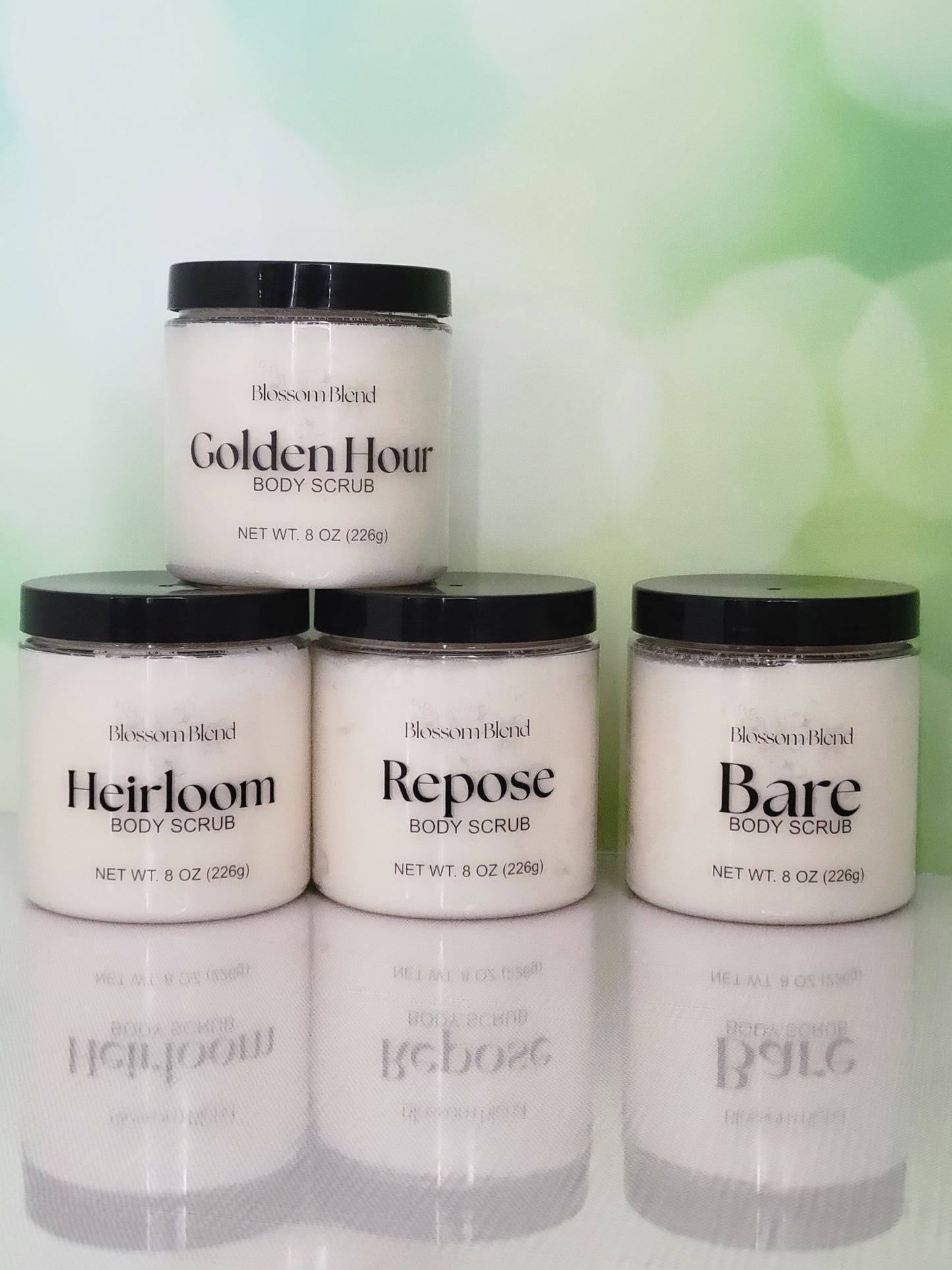 Five jars of Blossom Blend body scrubs on a reflective surface with a blurred green background.