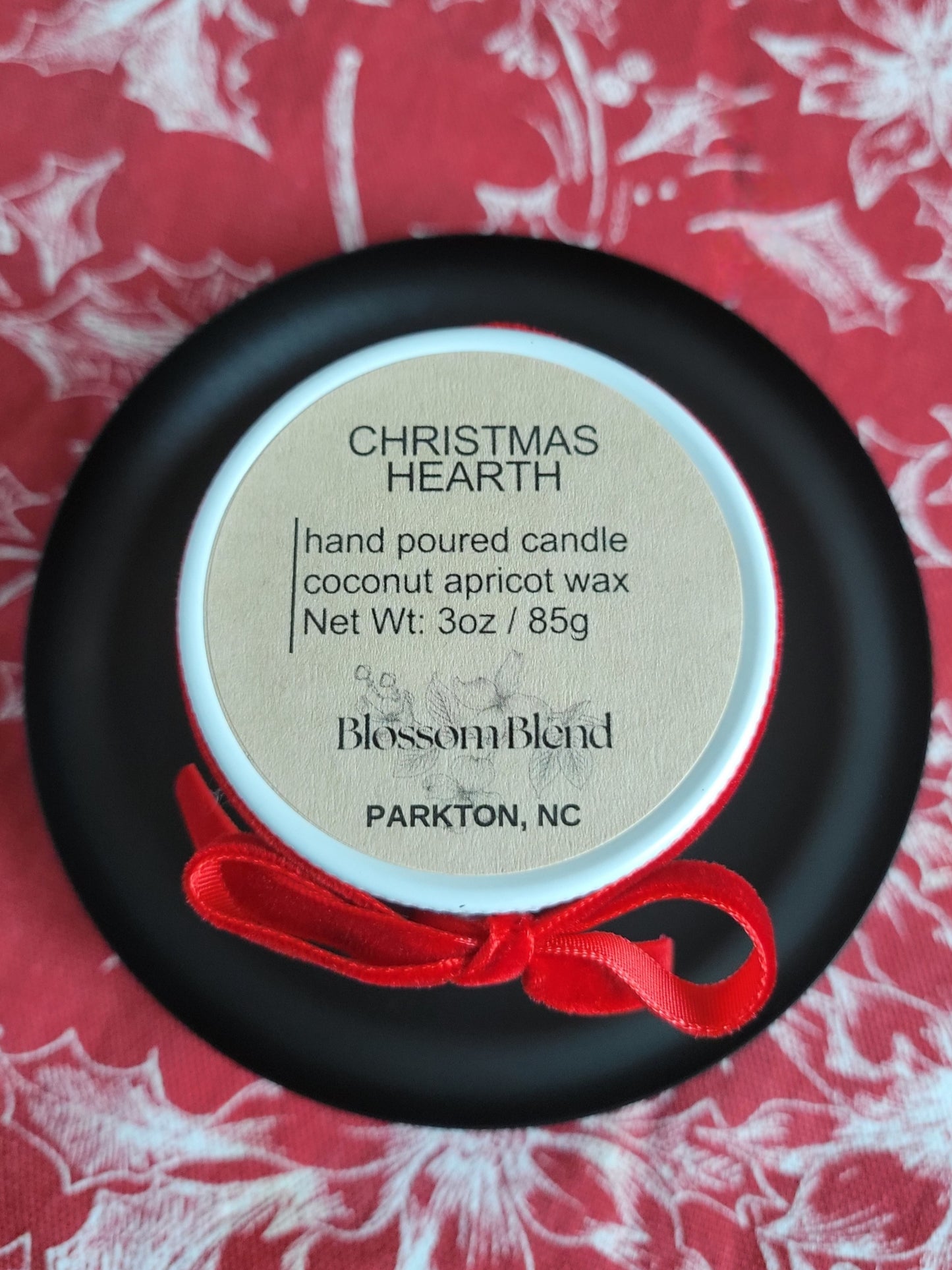 Hand poured candle labeled 'Christmas Hearth' by Blossom Blend on a red and white floral background.
