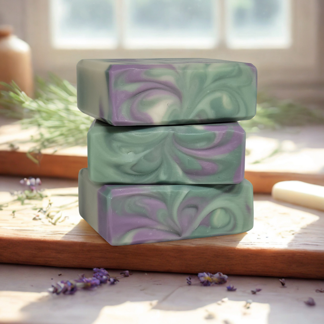 Three bars of green and purple soap stacked on a wooden surface with lavender flowers.
