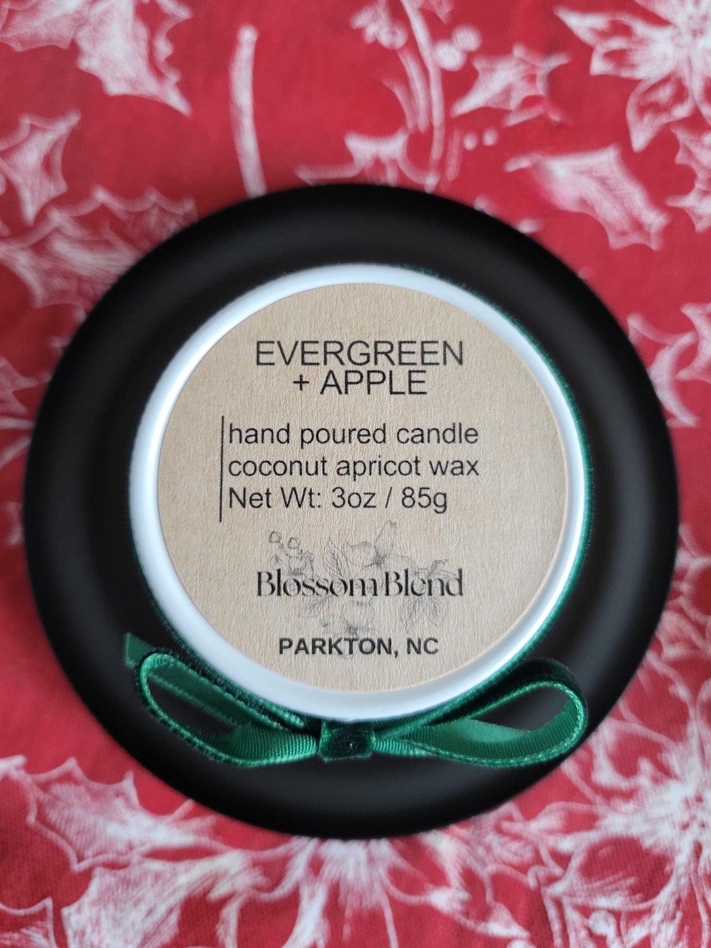 Hand poured candle labeled 'Evergreen + Apple' by Blossom Blend on a red floral background