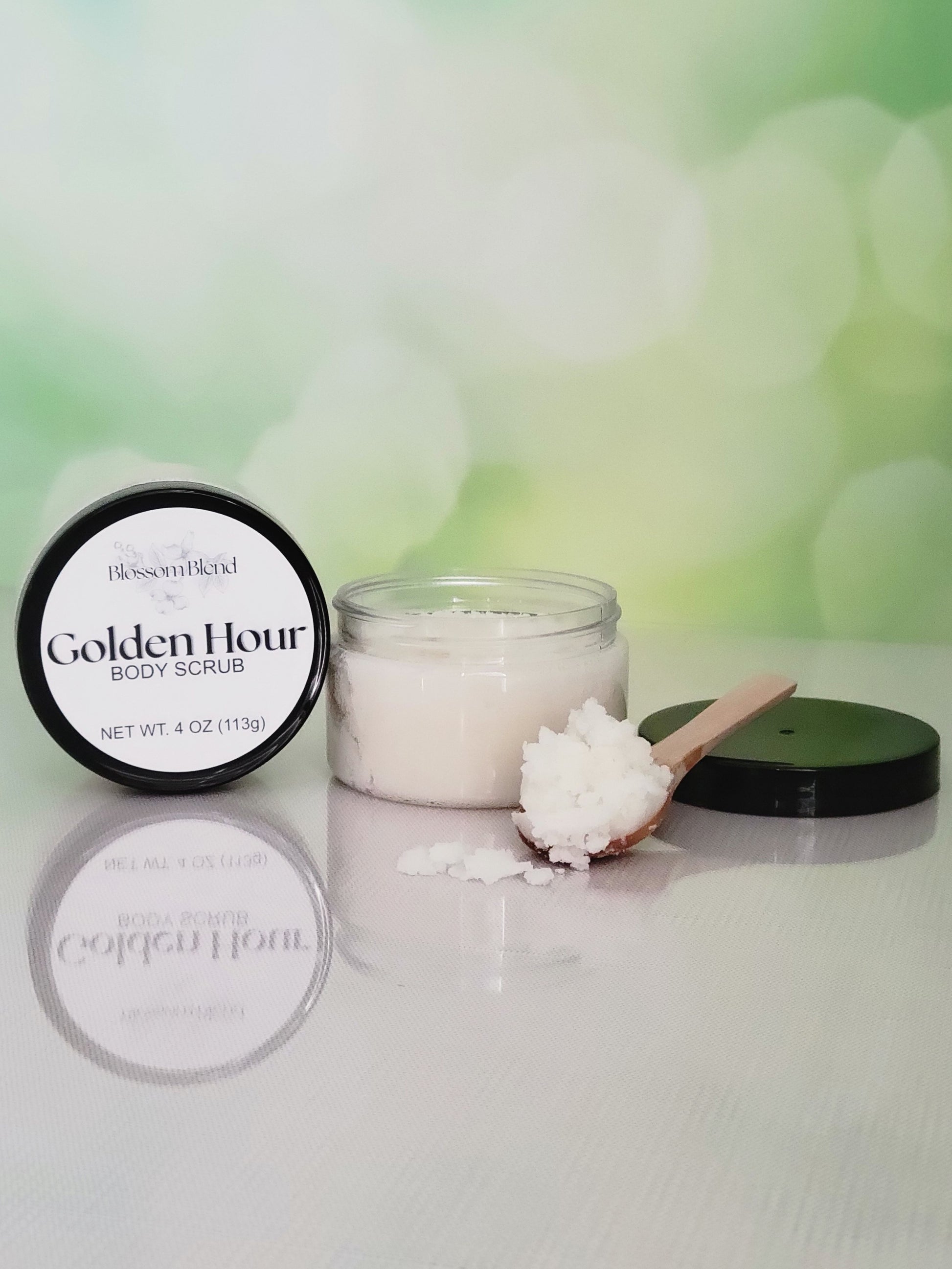 Golden Hour body scrub with a wooden scoop on a white surface with a green blurred background