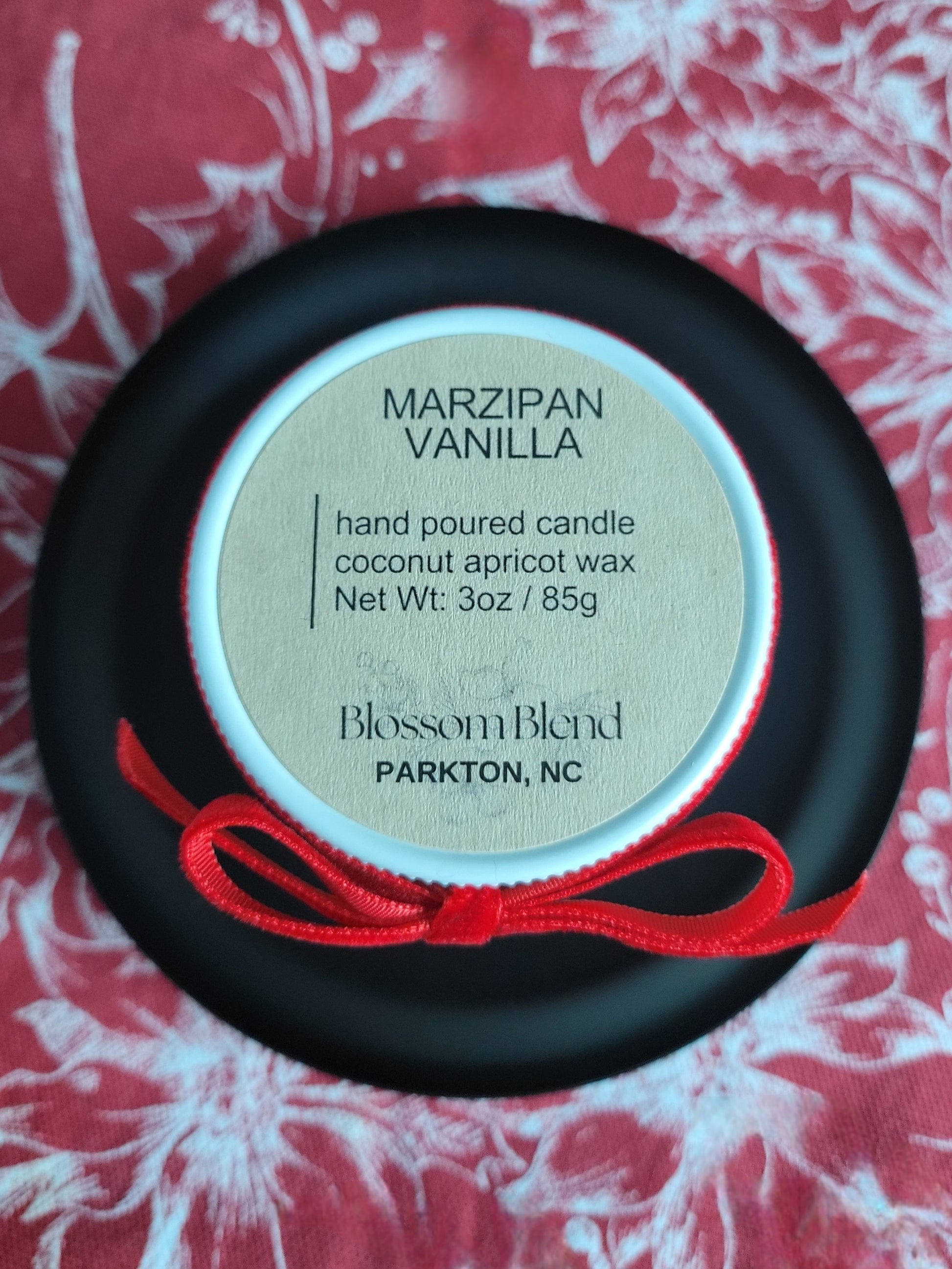 Marzipan Vanilla candle in a black container with a red string on a floral background