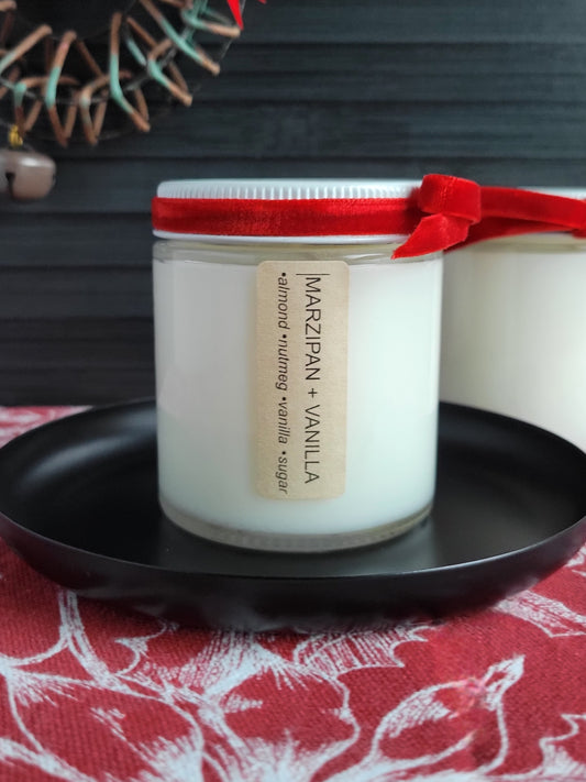 Candle jar labeled 'Marzipan + Vanilla' on a black plate with a red ribbon, against a dark background.