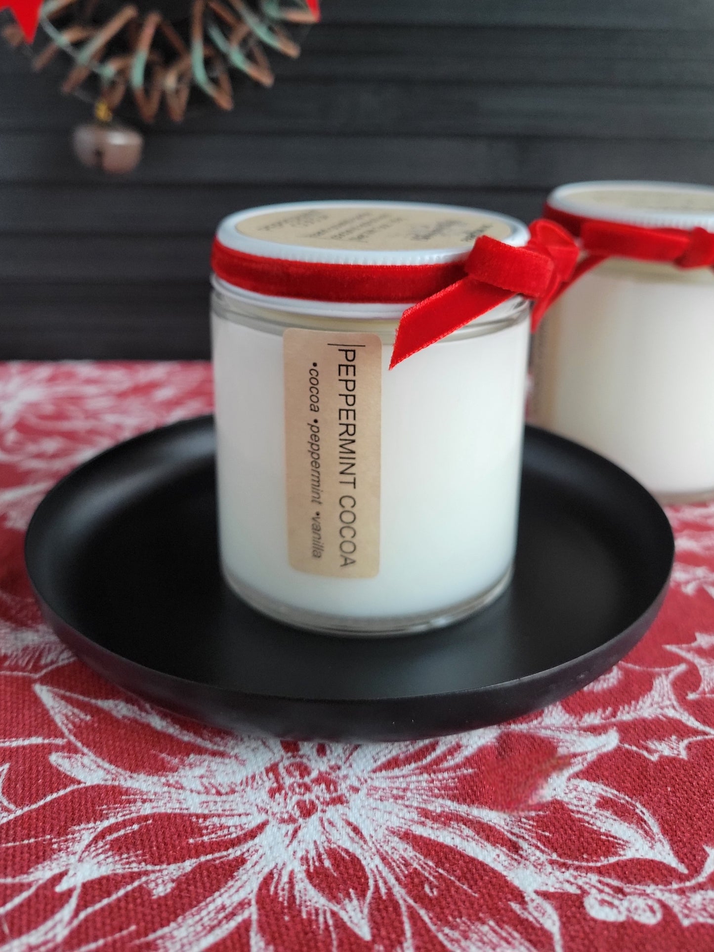 Candle jar labeled 'Peppermint Cocoa' with a red ribbon on a black plate, set against a patterned tablecloth.