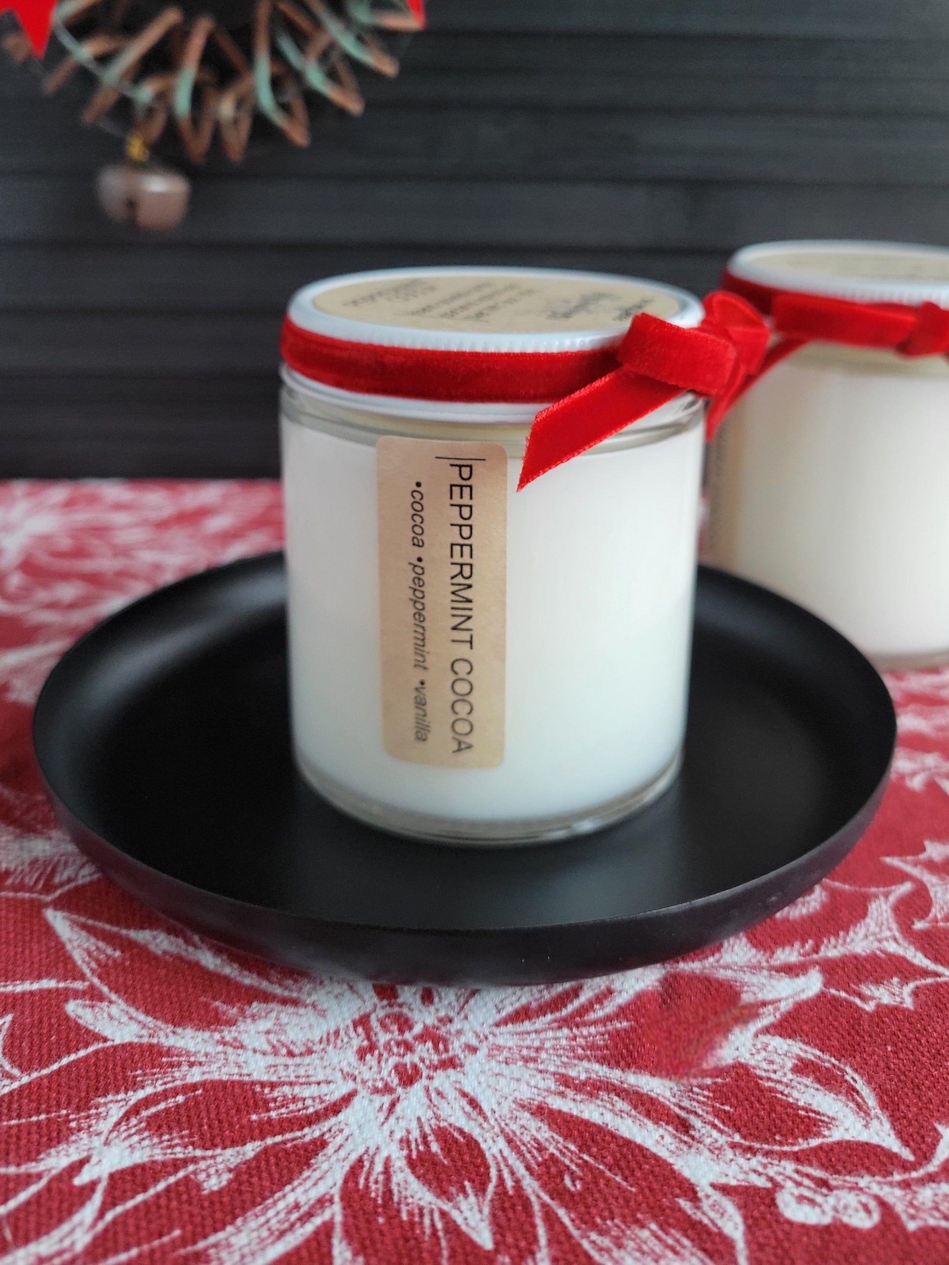 Candle jar labeled 'Peppermint Cocoa' with a red ribbon on a black plate, set against a patterned tablecloth.