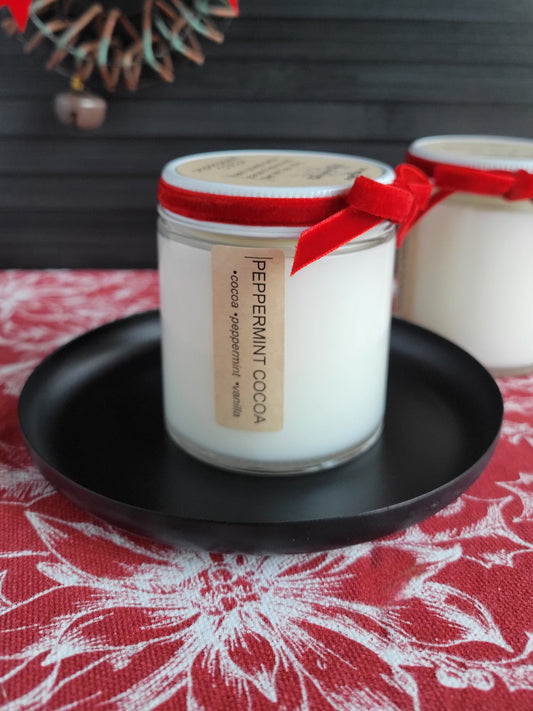 Candle jar labeled 'Peppermint Cocoa' with a red ribbon on a black plate, set against a patterned tablecloth.