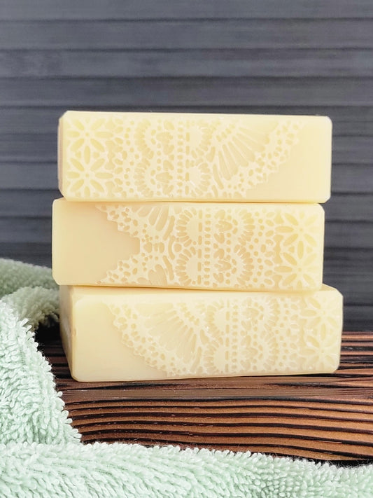 Three bars of textured soap stacked on a wooden surface with a towel underneath, against a gray background.