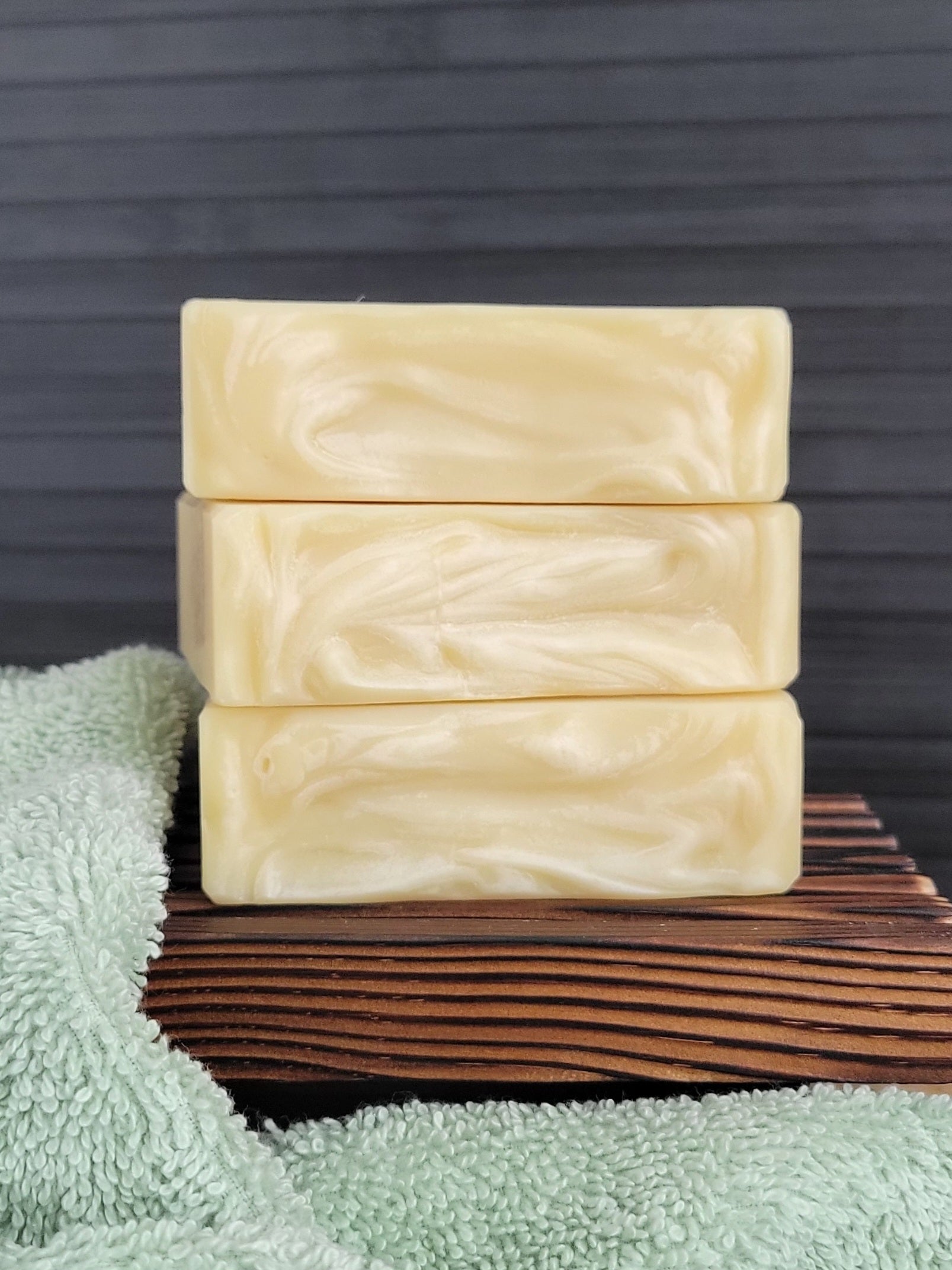 Three bars of soap stacked on a wooden block with a green towel underneath, against a dark background.